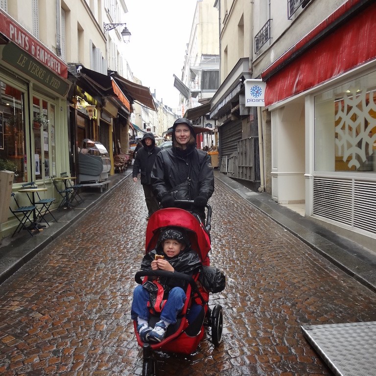 Boy eating an eclair in a stroller while mom pushes him through the rain.